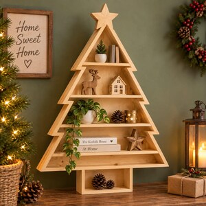 May include: A light-coloured wooden shelf shaped like a Christmas tree with a star topper. It features five horizontal tiers displaying a small potted plant, a wooden deer figurine, a miniature house, a trailing green plant, books, and pinecones.