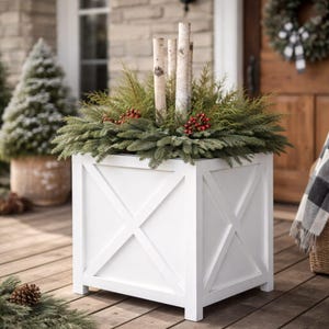 Peut inclure: Un bac à fleurs en bois blanc avec un motif en X sur chaque côté. La jardinière est remplie de branches de conifères, de baies rouges et de rondins de bouleau. Un élément décoratif pour les espaces extérieurs.