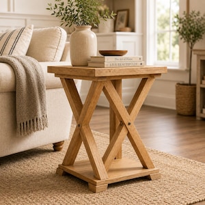 Può includere: Un tavolino quadrato in legno caratterizzato da una base a forma di X con quattro gambe e un ripiano inferiore. Il tavolo presenta una finitura naturale in venature di legno marrone chiaro ed è sormontato da un vaso in ceramica, una ciotola e due libri.
