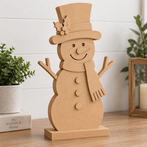 May include: A light brown, flat-cut wooden snowman decoration standing on a rectangular base. The snowman features a top hat with holly leaf and berry accents, a scarf, three circular buttons, and stick-like arms, all in a uniform matte finish.