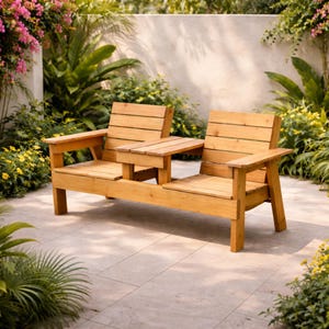 May include: A two-person wooden outdoor bench with a small table in the middle. The bench is made of light-colored wood and has a slatted back and seat. The bench is surrounded by lush greenery and flowers.