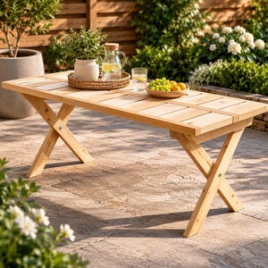 May include: A rectangular light-colored wooden outdoor table featuring a slatted top and crossed X-shaped legs. The table surface holds a round woven tray with a potted green plant, a glass pitcher of water with lemon slices, and a bowl of grapes.