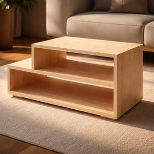 May include: A light-colored wooden coffee table with two shelves. The table has a rectangular shape with a stepped design, featuring a top section and a lower, extended section. The wood grain is visible, and the table sits on a neutral-colored rug.