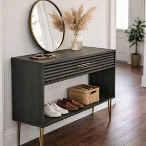 May include: A dark gray wooden console table with a drawer and a lower shelf. The table has gold-colored legs and a gold-framed mirror above it. A vase of dried flowers and a woven basket are on the table. Shoes are on the shelf.
