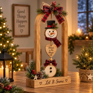 May include: A wooden arch tabletop decoration featuring a hanging snowman made of wood slices, a heart-shaped plaque with the text Believe in the Magic, and a base with the text Let It Snow. It is adorned with red and black plaid bows and pine.