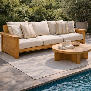 Puede incluir: Un sofá de exterior de madera clara con cojines beige y cojines decorativos. Una mesa de centro redonda de madera se encuentra frente al sofá, con dos tazas y un cuenco de frutos secos. Una alfombra de color claro está debajo del sofá y la mesa.