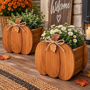 May include: Two wooden pumpkin-shaped planters with a natural brown finish. Each planter features a jute twine bow tied around the stem with a small wooden leaf cutout attached. The planters are filled with green foliage and white flowers.