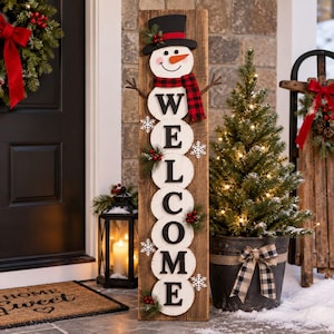 May include: A tall, rectangular wooden sign featuring a snowman at the top wearing a black top hat and a red and black plaid scarf. The sign displays the word WELCOME vertically in black capital letters on white circular wooden cutouts.