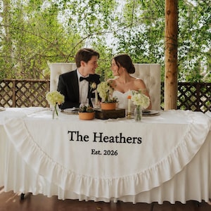 Może przedstawiać: Biały bieżnik ślubny z falbanką, ozdobiony czarnym haftowanym napisem The Heathers oraz Est. 2026. Bieżnik jest ułożony na białym obrusie i udekorowany małymi roślinami w doniczkach oraz białymi kompozycjami kwiatowymi.