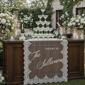 Puede incluir: Una barra de madera oscura está lista para una celebración, con una torre de copas de champán como pieza central. Un camino de mesa transparente con ribete de encaje muestra el texto "Cheers to The Sullivans". Arreglos florales blancos y velas completan el ambiente elegante.