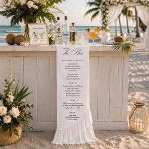 May include: A white fabric bar menu sign with a ruffled edge, featuring black printed text listing signature cocktails, spirits, wine, and beer. The sign hangs over a wooden bar on a beach, accompanied by liquor bottles and floral arrangements.