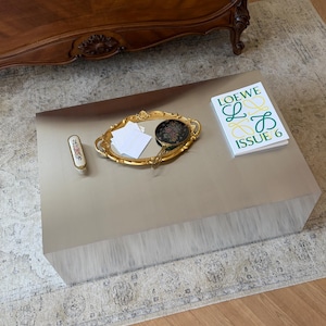 Könnte beinhalten: Ein goldfarbener rechteckiger Couchtisch mit einem dekorativen Tablett, einem kleinen Pinsel und einem Buch. Das Tablett enthält einen Kompaktspiegel und weiße Quadrate. Das Buch trägt den Text "LOEWE L B ISSUE 6". Ein Holzbetthaupt ist im Hintergrund.