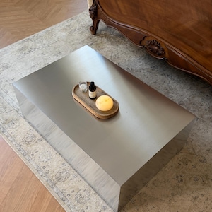 May include: A large, rectangular, brushed metal coffee table. A wooden tray on top holds a small glass, a dark bottle, and a yellow sphere. The table sits on a patterned rug, with a wooden bed frame in the background.
