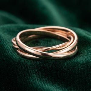 May include: A rose gold ring composed of three interlocking, smooth, polished bands. The metal has a warm, pinkish hue and a reflective surface, creating a continuous, intertwined circular structure that sits as a single cohesive piece of jewelry.