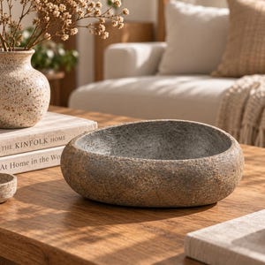 Puede incluir: Un cuenco de piedra bajo y redondeado con un exterior rugoso y texturizado y un interior gris liso. El cuenco tiene una forma orgánica similar a un guijarro y un aspecto natural y terroso, con una paleta de colores moteados en marrón y gris.