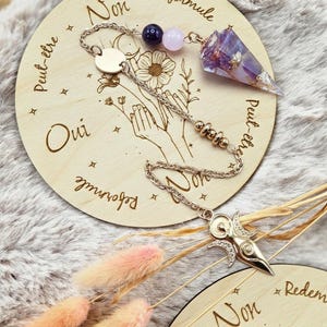 Op de afbeelding: Een waarzeggerijpendel met een paarse kristallen punt en zilveren ketting, rustend op een houten schijf met de woorden "Oui" en "Non" gegraveerd. De pendel heeft een zilveren bedel aan het uiteinde en is versierd met kralen.