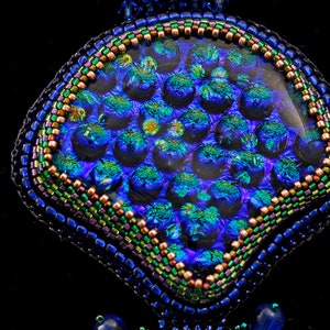 May include: A close-up of a blue, green, and purple beaded pendant with a round, iridescent glass center. The pendant is surrounded by a beaded border with a variety of colors.