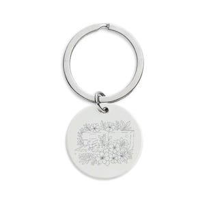 Puede incluir: Un llavero plateado con un colgante redondo y blanco. El colgante presenta un dibujo lineal de una caravana rodeada de flores y follaje. El anillo del llavero está unido al colgante con un pequeño lazo.