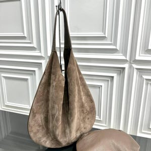 May include: A large, slouchy hobo bag crafted from brown suede with a single integrated shoulder strap. Beside it sits a smaller, rounded brown leather pouch featuring a gold-toned zipper closure and a delicate gold chain strap attached to the side.