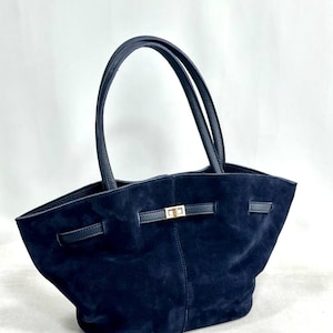 May include: A dark navy blue suede tote bag featuring two tall shoulder straps, a central gold-tone metal turn-lock closure, and a horizontal strap detail across the front. The bag has a wide, open-top design with a soft, textured finish.