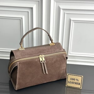 May include: A brown suede handbag with a top handle and a gold-toned zip closure featuring two pulls. A small rectangular sign next to the bag displays the text 100% LEATHER in black lettering on a gold background, confirming the material.