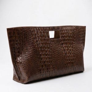 Könnte beinhalten: Braune, rechteckige Clutch-Tasche mit Krokodilleder-Textur. Die Tasche hat einen rechteckigen Griffausschnitt und eine breite Öffnung. Die Tasche ist aus glänzendem Kunstleder gefertigt.