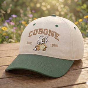 May include: A cream-colored baseball cap with a dark green brim and button top. The front features an embroidered graphic of the character Cubone, the text CUBONE, and the text EST. 1996 in brown lettering. The cap has dark green eyelet vents.