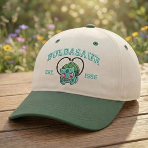 May include: A baseball cap with a white crown and a green peak. The cap features the word "Bulbasaur" embroidered in teal, along with a Bulbasaur graphic and the text "EST. 1996". The cap has a green button on top.