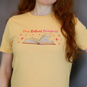May include: A light yellow t-shirt with the text "Fiery Redhead Protagonist" above an open book illustration. The design is in red and the shirt is short-sleeved.