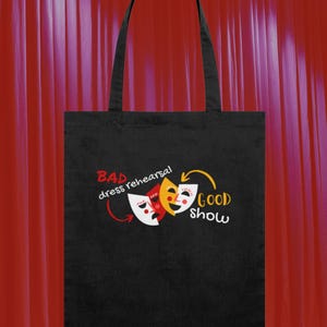 May include: Black tote bag with a theatre-themed design. It features three masks in red, yellow, and white, with the text "BAD dress rehearsal" and "GOOD show". The bag has two black handles.