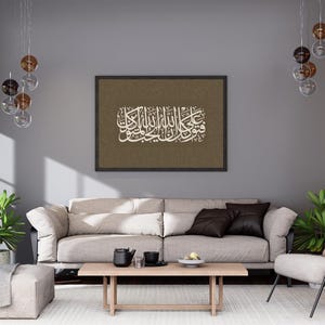 Könnte beinhalten: Ein gerahmtes Kunstwerk mit weißer arabischer Kalligraphie auf einem strukturierten braunen Hintergrund. Das Kunstwerk wird über einem hellfarbigen Sofa mit dunkelbraunen Zierkissen gezeigt. Der Raum hat ein modernes Design mit grauen Wänden und dekorativer Beleuchtung.