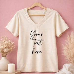 May include: Cream-colored V-neck t-shirt with the text "Your Text Here" in black script. The shirt is hanging on a wooden hanger against a pink background. Dried flowers and a candle are in the foreground.