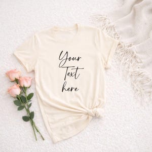 Op de afbeelding: Crèmekleurig T-shirt met de tekst "Your Text here" in een zwart scriptlettertype. Het T-shirt is aan de onderkant geknoopt en ligt naast een boeket roze rozen. Het shirt ligt op een wit, getextureerd oppervlak.