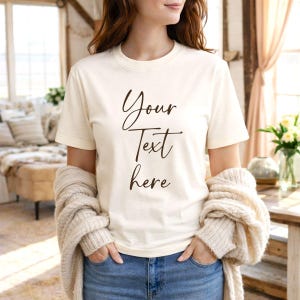 May include: Cream-colored t-shirt with the text "Your Text here" in a dark brown script. The model is wearing blue jeans and a beige sweater. The shirt is a crew neck style.
