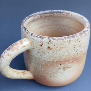 May include: A speckled beige ceramic mug with a rounded handle. The mug has a light brown glaze with darker brown speckles.