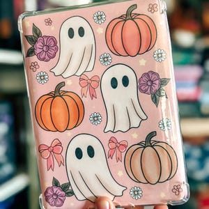 Peut inclure: Une coque de protection transparente pour tablette avec un fond rose clair orné d'un motif répétitif de fantômes blancs, de citrouilles orange, de fleurs violettes et de petits nœuds roses, agrémenté de minuscules étoiles blanches.