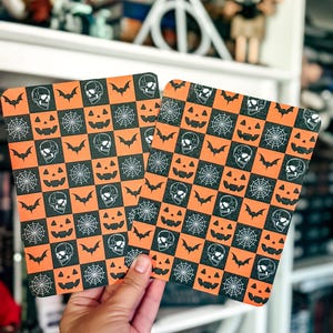 May include: Two rectangular flat items featuring a repeating checkerboard pattern of orange and black squares. Each square contains a white line-art illustration of a skull, a bat, a spider web, or a jack-o'-lantern face in a grid layout.