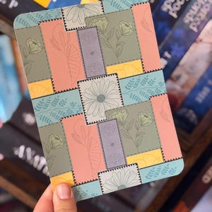 Puede incluir: Un posavasos rectangular con un dise&ntilde;o de patchwork que presenta ilustraciones florales y de hojas en colores pastel. El posavasos tiene una combinaci&oacute;n de colores azul claro, rosa, amarillo y verde, con un borde gris y una margarita blanca.