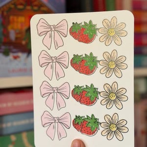 Może przedstawiać: Biała karta z ilustracjami różowych kokard, czerwonych truskawek z zielonymi liśćmi i kremowych stokrotek z żółtymi środkami. Karta jest trzymana w dłoni, z rozmytym tłem książek.