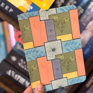 Puede incluir: Un cuaderno rectangular con un dise&ntilde;o floral de patchwork. El dise&ntilde;o presenta varios colores, incluyendo naranja, verde, azul y amarillo, con ilustraciones florales y de hojas. El cuaderno est&aacute; en la mano de una persona.