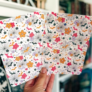 May include: Two rectangular sheets featuring a repeating pattern of black spiderwebs, orange and pink flowers, and small purple and orange bats on a white background. One sheet has a smooth finish, while the other has a holographic glitter overlay.