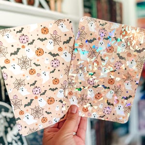 May include: Two rectangular sheets featuring a repeating Halloween pattern of white ghosts, orange and pink pumpkins, black bats, and spiderwebs. One sheet has a matte finish, while the other is overlaid with a reflective, holographic shard effect.