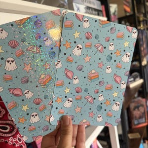 Puede incluir: Dos tarjetas rectangulares con fondo azul claro que presentan un patrón repetitivo de fantasmas blancos con ojos de corazón, conchas rosas, estrellas de mar naranjas, pilas de libros y pequeños paraguas. Una tarjeta tiene un acabado holográfico.