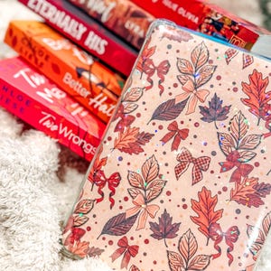 May include: A rectangular tablet case featuring a light peach background with a repeating pattern of autumn leaves, small red bows, and scattered dots. The surface has a holographic finish that reflects light with small star-shaped sparkles.