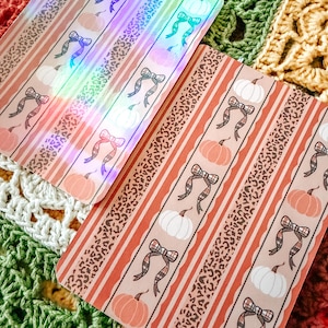Op de afbeelding: Twee rechthoekige stickers met een herhalend patroon van oranje en witte pompoenen, geruite strikken en luipaardprint strepen. Eén sticker heeft een holografische afwerking met regenboogreflecties, de andere heeft een matte afwerking.