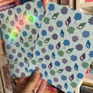 Puede incluir: Dos tarjetas rectangulares con esquinas redondeadas que presentan un fondo azul claro con un patrón de varias conchas marinas en tonos azul oscuro, morado y verde, intercaladas con pequeños puntos blancos y lazos finos de color verde claro.