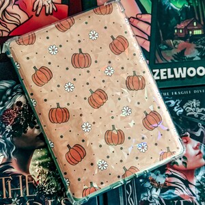 Puede incluir: Una funda protectora rectangular transparente para lector electrónico o tableta con un patrón repetitivo de calabazas naranjas y pequeñas margaritas blancas sobre un fondo bronceado claro con un sutil efecto geométrico holográfico.