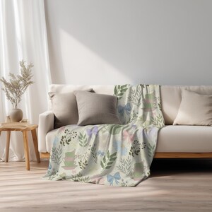 Puede incluir: Una manta de color beige claro sobre un sofá, con un patrón repetido de tazas de té, lazos y ramitas de hojas en colores pastel. El diseño de la manta incluye elementos rosas, azules y verdes, creando una estética suave y caprichosa.