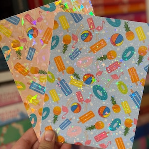 May include: Two sheets of paper with a summer-themed pattern. One sheet has a light pink background, the other is light blue. Both feature holographic accents and illustrations of popsicles, pineapples, beach balls, and flamingos.