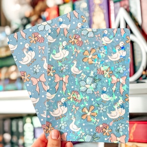 May include: Two light blue cards featuring a pattern of white ducks, pink bows, and flowers. The cards have a holographic finish. The cards are held in front of a blurred background of books and decorations.